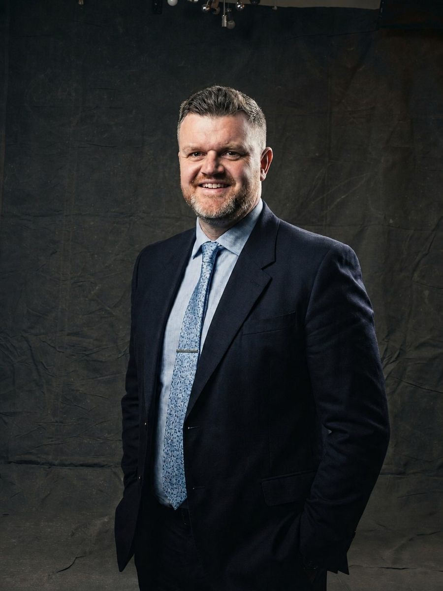 Krystian Liptrot smiling, in a navy suit against a dark backdrop